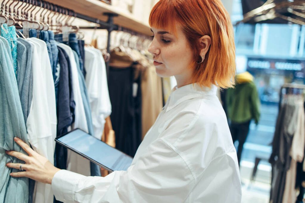 Woman with shoulder length, straight orange hair looking through a rack of clothes and making notes on an iPad.