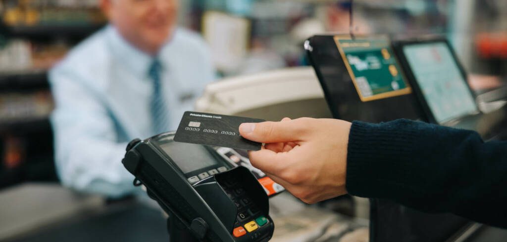 A hand holding a credit card to tap and pay at a cash register of a convenience store.