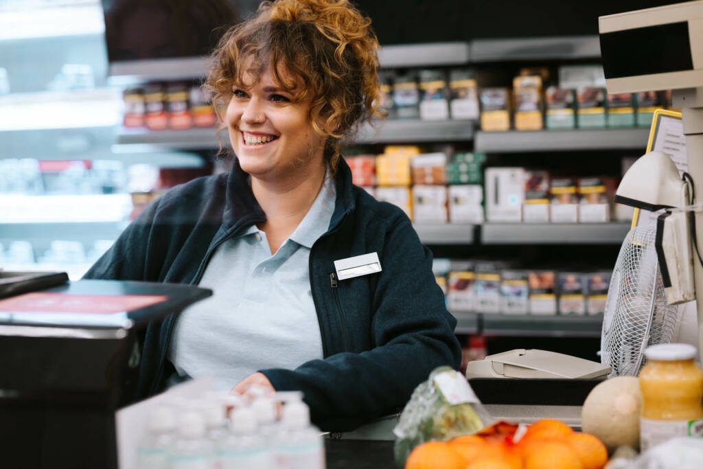 Woman with curly brown hair in ponytail smiles as she works behind a cash register of a convenience store.