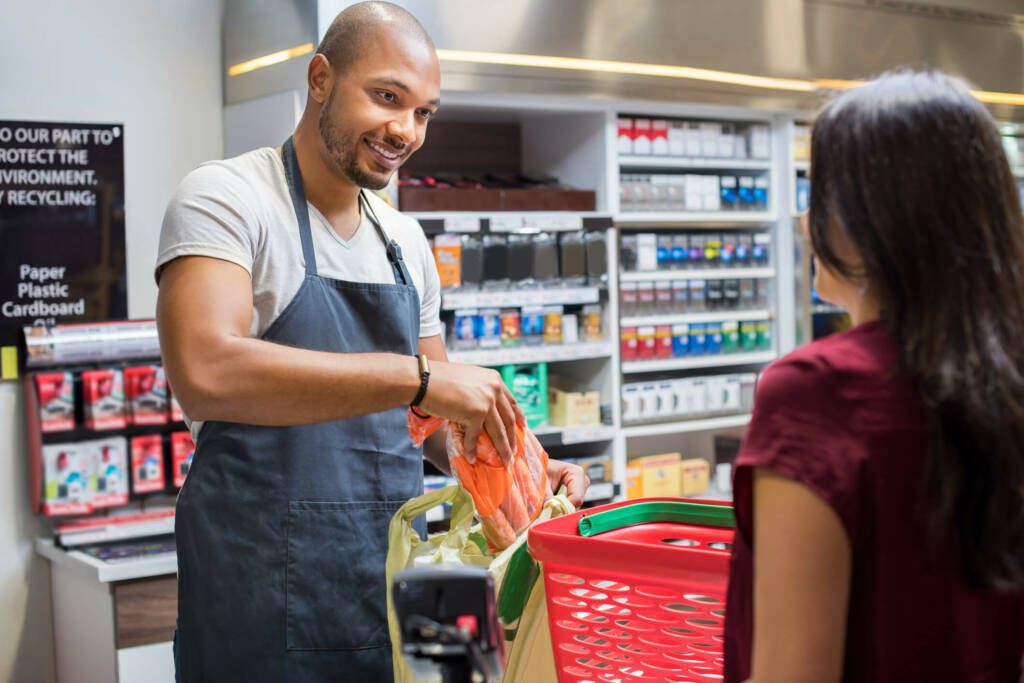 Male convenience store employee puts bags groceries while chatting with customer