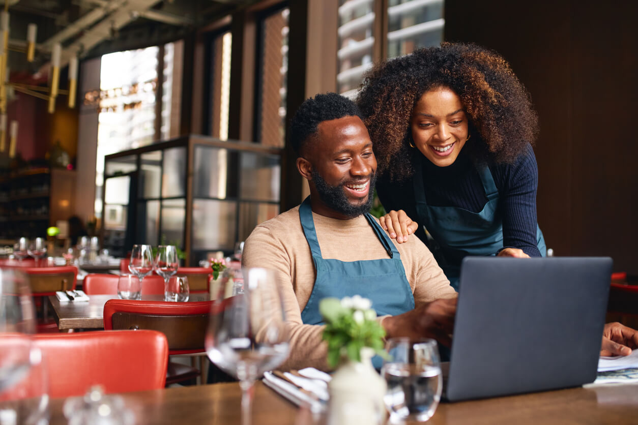 An African American man and woman in a restaurant are sitting at a table looking at information on a laptop together.